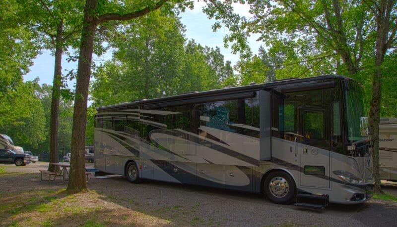 A large, modern RV is parked at a wooded campsite with a picnic table nearby and other RVs visible in the background.