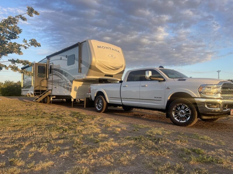 A white pickup truck is parked in front of a large Montana fifth-wheel RV trailer on a dry, grassy area under a partly cloudy sky at sunset.