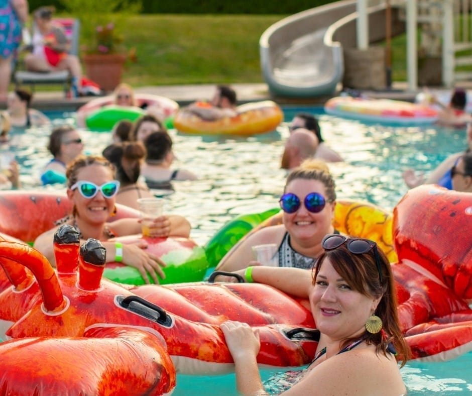 People relaxing on colorful pool floats at a crowded outdoor swimming pool, with some holding drinks and smiling at the camera.