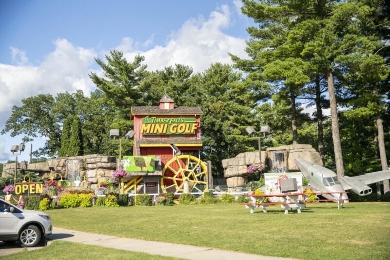 Timber Falls Mini Golf entrance with a large yellow sign, water wheel, plane display, picnic tables, flowers, and trees in the background.