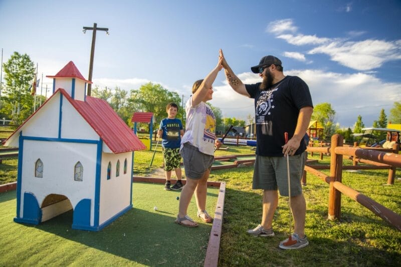 Two people high-five on a miniature golf course near a church-shaped obstacle, while a child stands in the background on a sunny day.