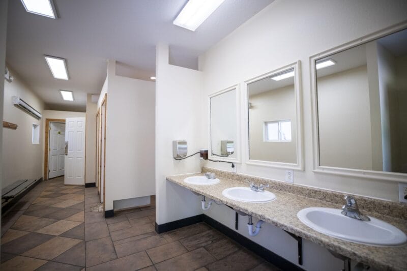 A clean public restroom with three sinks, large mirrors, and tiled floors under bright overhead lighting. A hallway and open stalls are visible in the background.