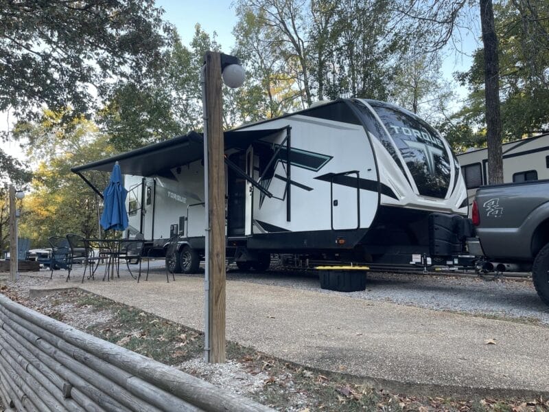 A large white travel trailer with an awning extended is parked at a campsite next to a pickup truck, with a patio area and outdoor table nearby, surrounded by trees.