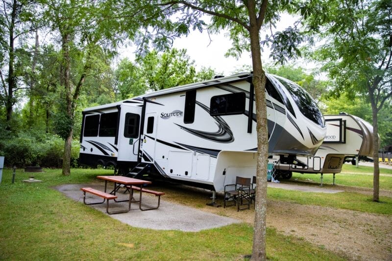 A large white and black fifth-wheel RV parked at a campsite with a picnic table and trees nearby.