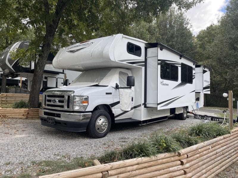 A white motorhome with extended slide-outs is parked on a gravel campsite under trees, with a wooden barrier and utility hookups nearby.