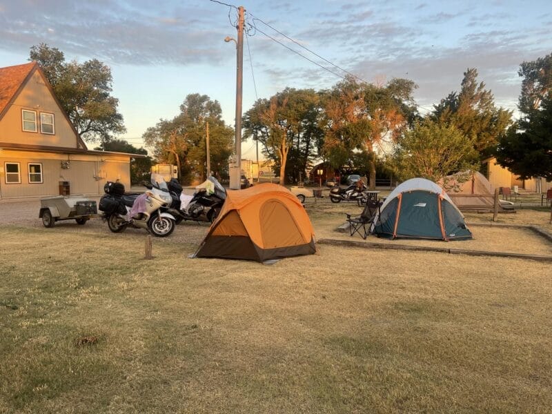 Two tents are set up on a grassy area near two motorcycles and a house, with camping chairs and equipment visible in the early evening light.