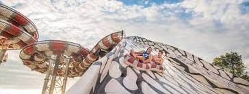 Two people ride a large inflatable raft down a patterned water slide at an outdoor water park under a partly cloudy sky.