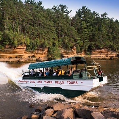 A tour boat labeled "LAND AND WATER DELLS TOURS" drives from land into a river, carrying passengers, with rocky cliffs and dense trees in the background.