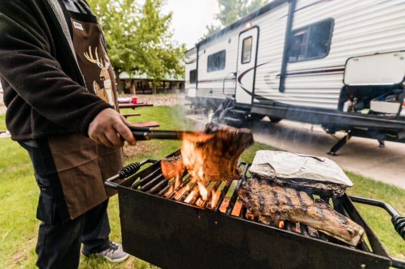 A person grills meat over an open flame on a portable grill outside, with a camper trailer and trees in the background.
