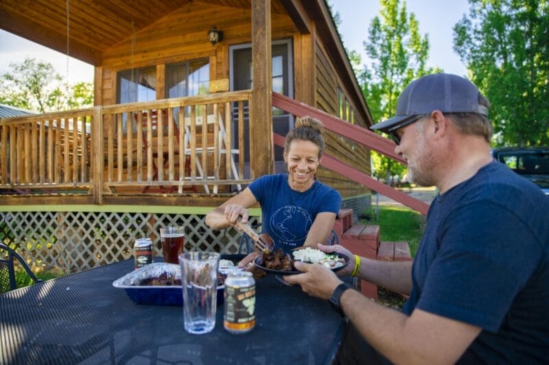 Two people sit at an outdoor table by a wooden cabin, serving and eating food with drinks, on a sunny day.