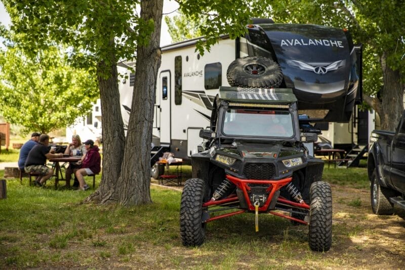 A black off-road vehicle is parked in front of a large Avalanche RV. Four people sit at a picnic table under trees, with camping items on the grass nearby.