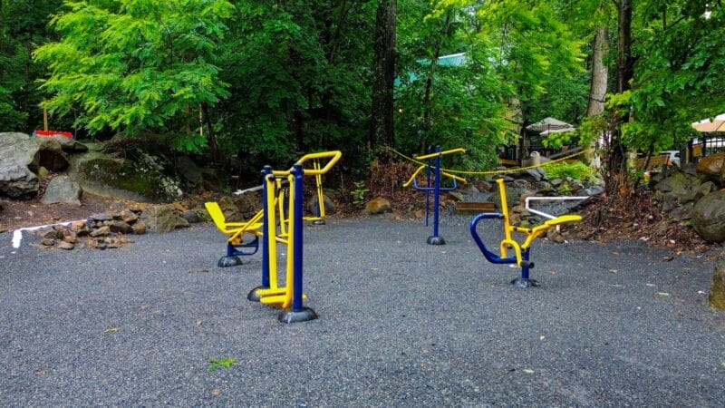 Outdoor fitness area with yellow and blue exercise equipment on a gravel surface, surrounded by trees and greenery.
