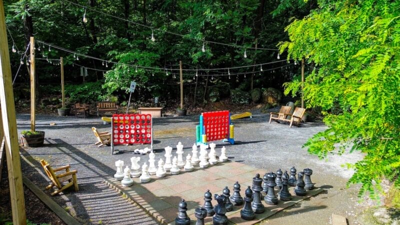 Outdoor game area with oversized chess set and two large Connect Four boards, surrounded by benches and string lights, with trees in the background.