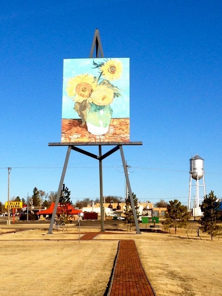 A large outdoor easel holds an oversized replica of Van Gogh's "Sunflowers" painting in a park, with a water tower and buildings in the background.