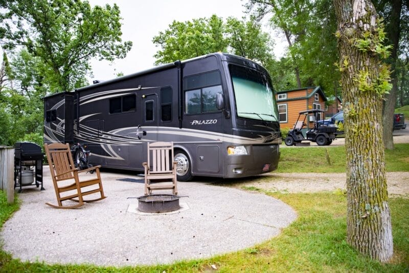 A large black RV is parked at a campsite near a small wooden cabin, with two wooden rocking chairs and a fire pit in the foreground.