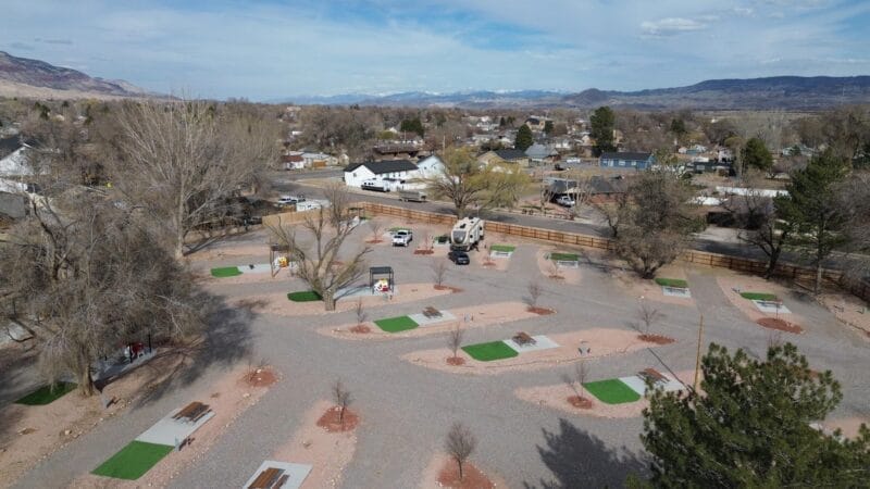 Aerial view of an RV park with empty gravel parking pads, picnic tables, a few parked vehicles, scattered trees, and nearby residential houses. Mountains are visible in the background.