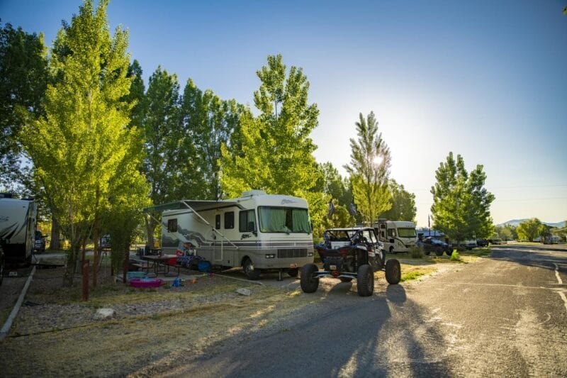 A parked RV at a campsite with trees, outdoor chairs, and a recreational off-road vehicle nearby under bright sunlight.