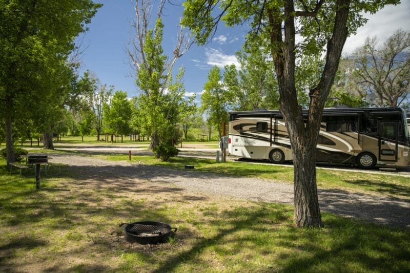 An RV is parked at a shaded campsite with picnic tables, a fire ring, and trees under a partly cloudy sky.