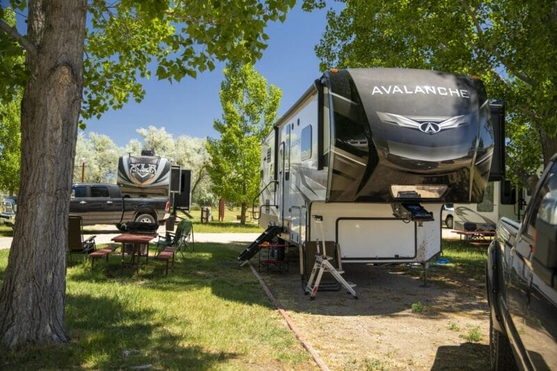 Two large RVs are parked at a campground under leafy trees, with picnic tables and chairs set up nearby on a sunny day.