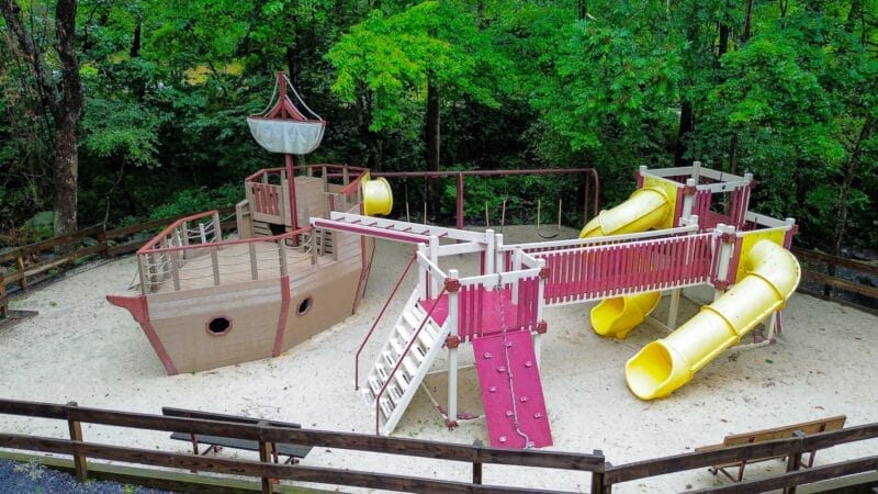 A wooden playground shaped like a pirate ship with slides, climbing walls, and ladders, surrounded by trees and enclosed by a fence.
