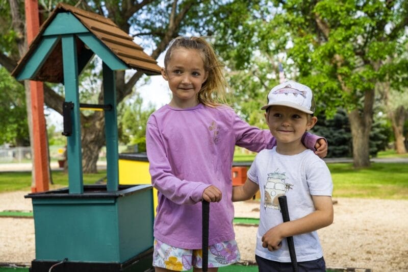 Two children stand side by side outdoors, holding mini golf putters, with one arm around each other near a small well structure on a mini golf course.