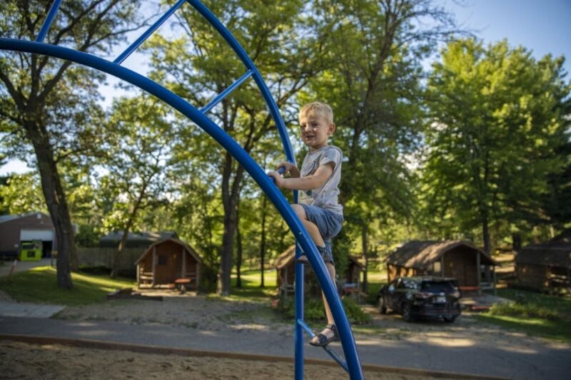 A young boy climbs on a blue metal playground structure, with trees, cabins, and parked vehicles in the background.