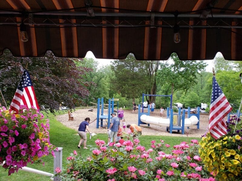 Children and adults gather near playground equipment in a park surrounded by trees, flowers, and two American flags under a striped awning.