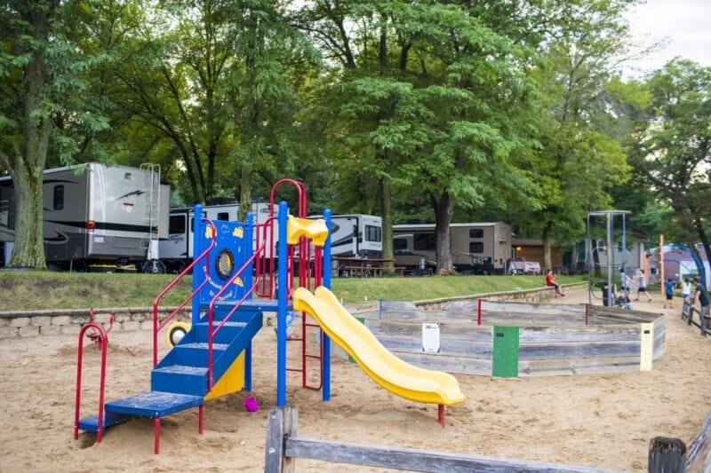 Playground with a blue and yellow slide, surrounded by sand, near RVs and trees in a park-like setting.