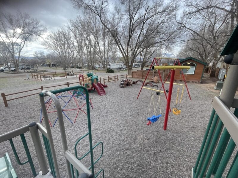 A playground with swings, a slide, climbing structures, and a playhouse, surrounded by leafless trees and wooden fences on a cloudy day.