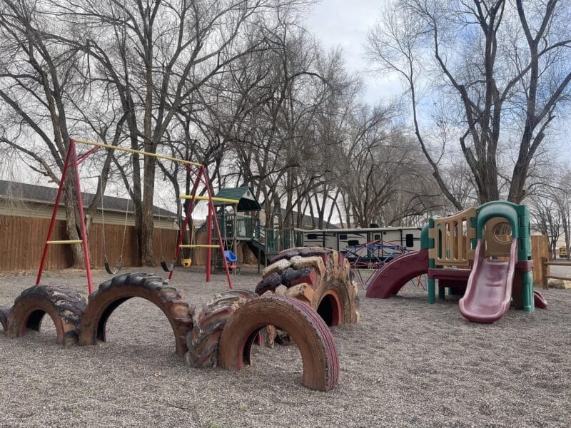 Outdoor playground with swings, a slide, climbing equipment, and large half-buried tires, surrounded by trees and a wooden fence under a cloudy sky.