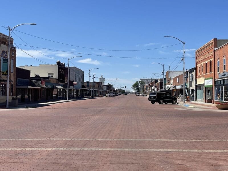 A wide, empty brick-paved street runs through a small town lined with low-rise storefronts under a clear blue sky.