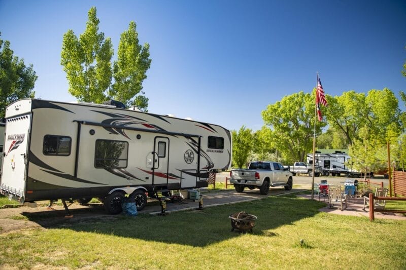 A travel trailer is parked on grass beside a pickup truck at a campground, with an American flag, picnic table, and trees in the background.
