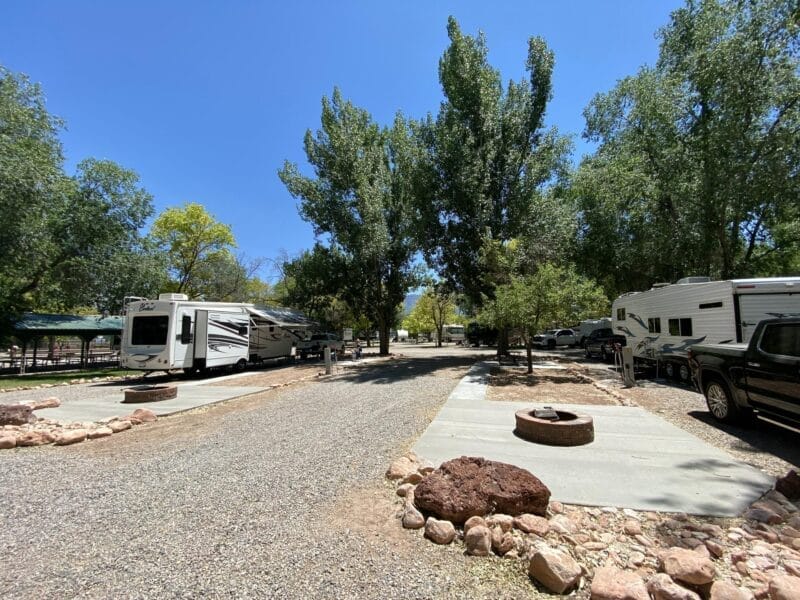 RV campground with several parked RVs, gravel roads, concrete pads, fire pits, and trees providing shade under a clear blue sky.