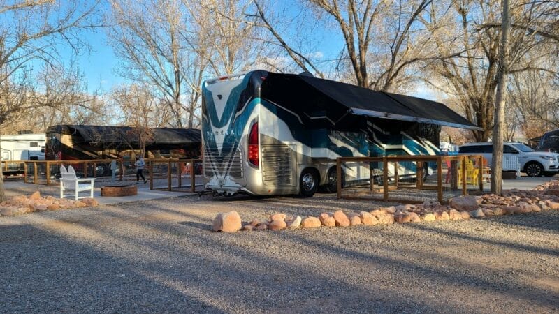 A large RV with extended awnings is parked at a campsite with a fire pit, two white chairs, and a gravel driveway, surrounded by leafless trees.