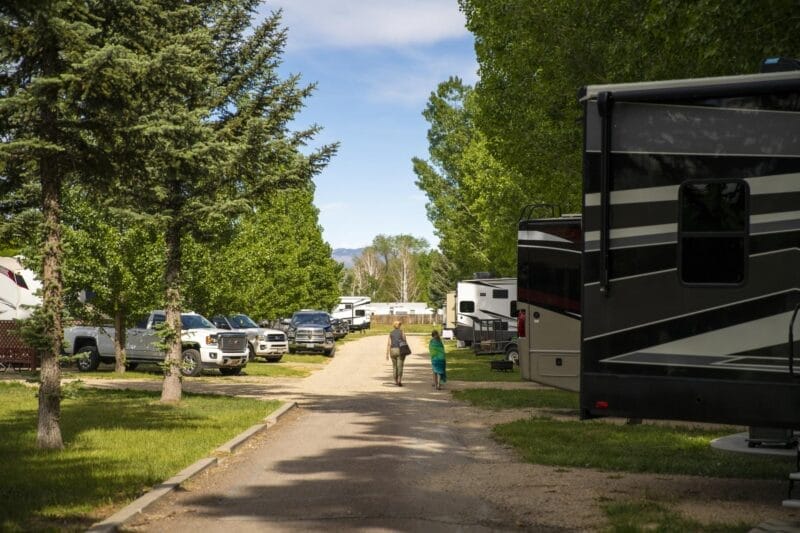 Two people walk down a gravel road in an RV park, surrounded by parked RVs, trucks, and green trees under a blue sky.