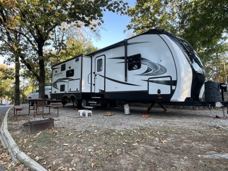 A large white and gray travel trailer is parked at a campsite with a picnic table, grill, and trees surrounding the area.