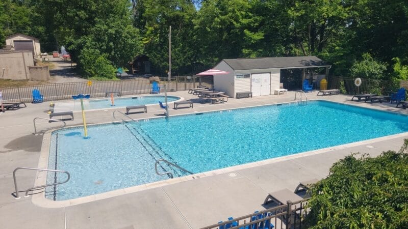 Outdoor rectangular swimming pool with clear water, surrounded by lounge chairs, tables, a small shed, and trees in the background, under a sunny sky.