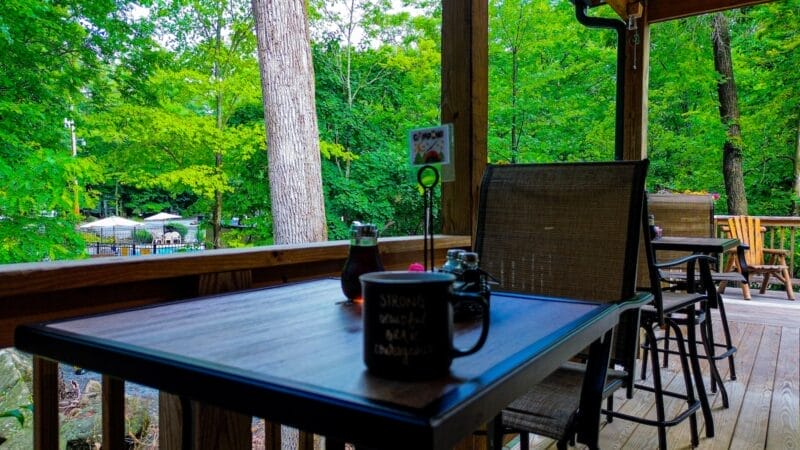 A shaded outdoor patio with a wooden table and chairs, surrounded by green trees and overlooking a pool area in the background.