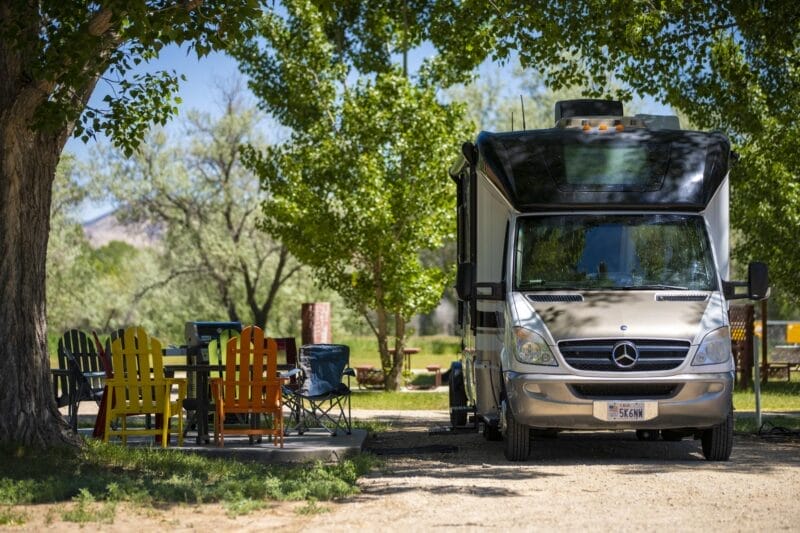 A large Mercedes-Benz RV is parked at a campsite with colorful Adirondack chairs and a table set up nearby, surrounded by trees in daylight.