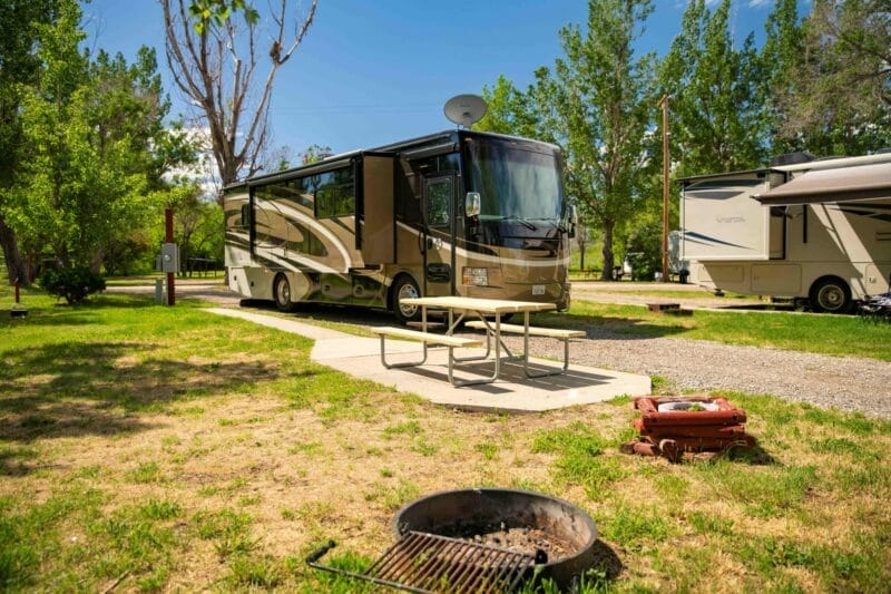 A large motorhome is parked at a campsite with a picnic table, a fire pit, and trees in the background on a sunny day.