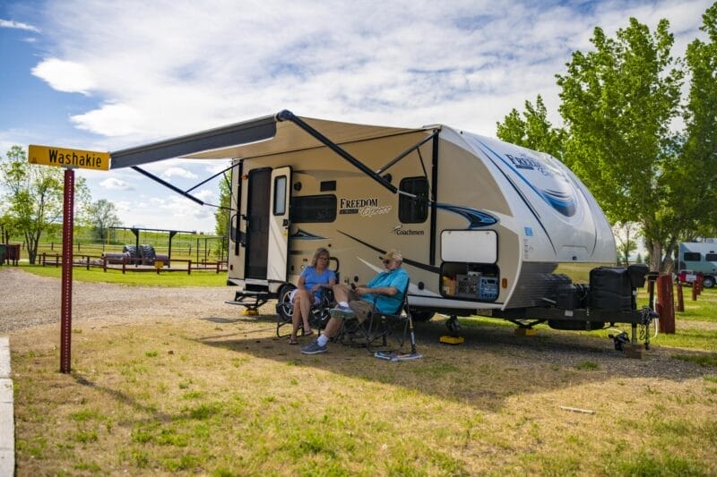 Two people sit in camping chairs under the awning of a travel trailer at a campsite marked "Washakie," with trees and other RVs in the background.