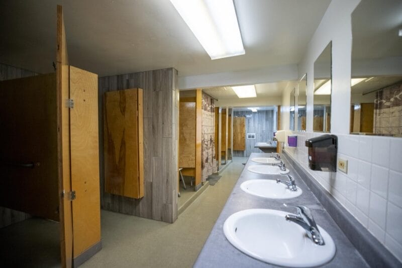 A public restroom with multiple sinks, mirrors, soap dispensers, and several wooden stall doors along the left side.