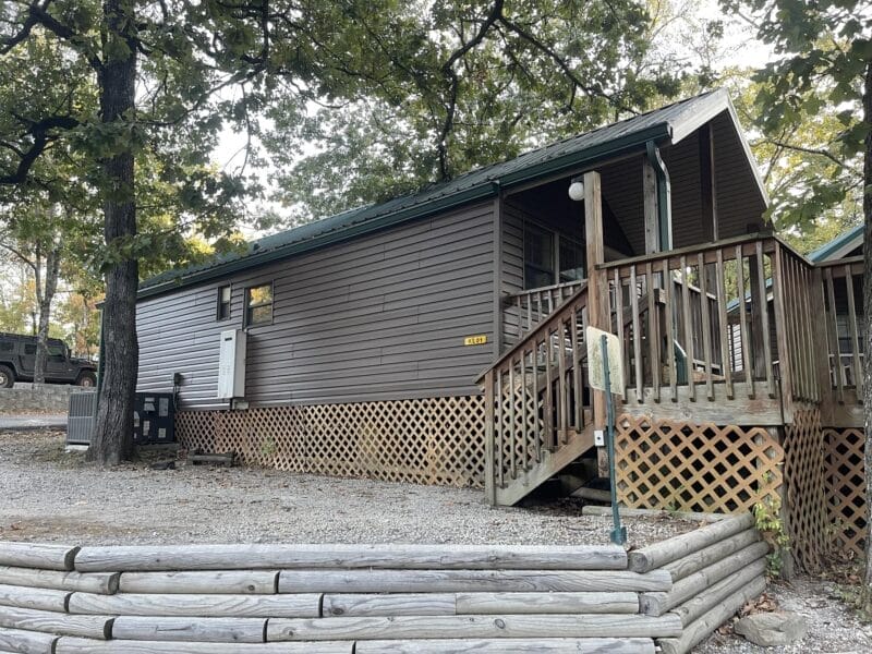 A brown wooden cabin with a green roof, elevated on a lattice foundation, with stairs leading to a small covered porch. Trees and gravel surround the building.
