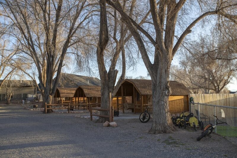 Row of wooden cabins among leafless trees, with several bicycles parked near a fence and a gravel path in the foreground.