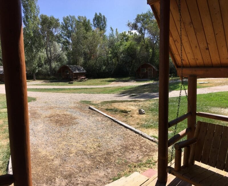 View from a wooden porch with a swing, overlooking a gravel path and several small log cabins surrounded by grass and trees.