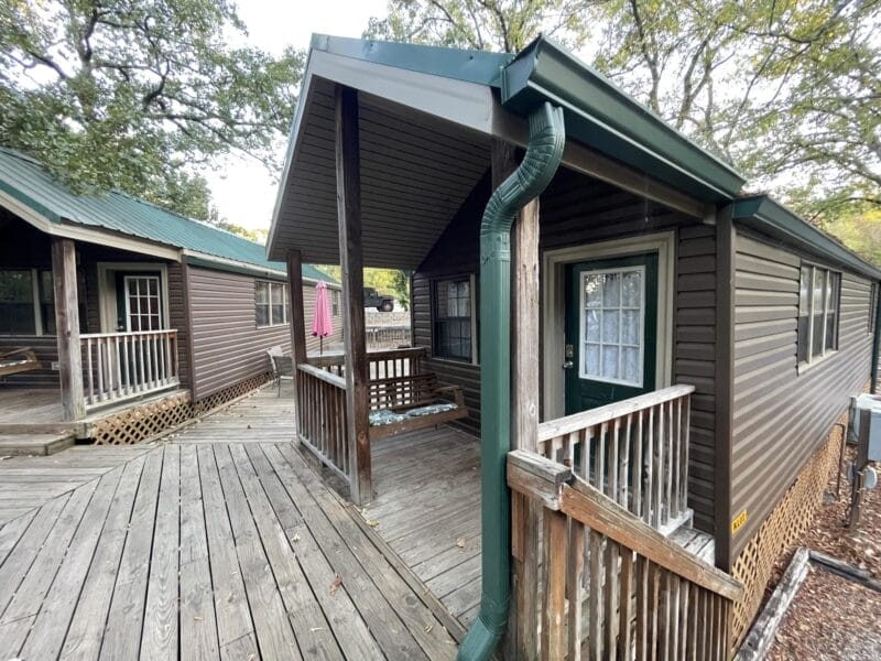 Two small brown cabins with green roofs are connected by a wooden ramp and surrounded by trees. One cabin features a small covered porch with railing.