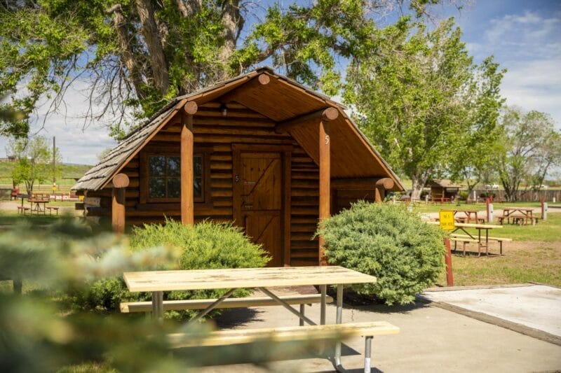 A small wooden cabin with a porch sits among trees and bushes, with a picnic table and benches on a concrete pad in front.