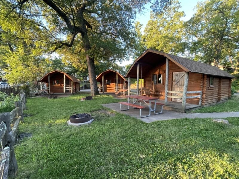 Three small wooden cabins are arranged on a grassy lawn with a picnic table and fire pit in front, surrounded by trees and a wooden fence.