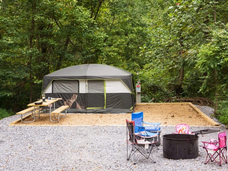 A large gray tent is set up on a sandy pad in a wooded area, with a picnic table, three folding chairs, and a fire pit in front.