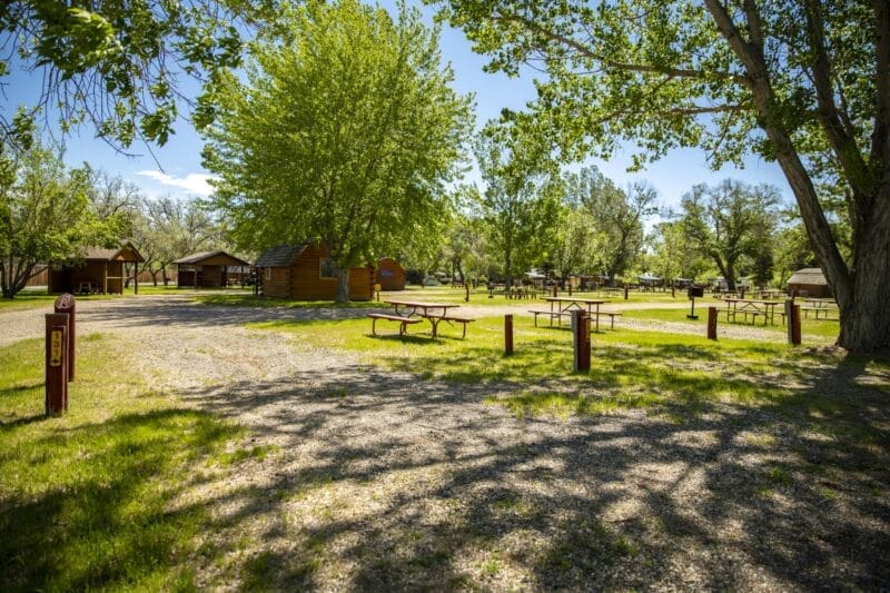 A sunny campground with picnic tables, gravel lots, grassy areas, and trees providing shade. Several small cabins and wooden shelters are visible in the background.
